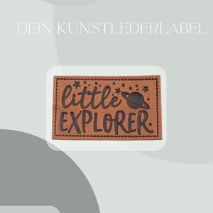 Könnte beinhalten: Ein braunes Lederpatch mit den Worten "little EXPLORER" in schwarzer Schrift. Das Patch zeigt ein Planeten- und Sternendesign. Der Text "DEIN KUNSTLEDERLABEL" steht oben und "Inklusive Anleitung und Test-Bügelbild" unten.