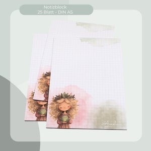May include: Three A5-sized notepads with a grid pattern and a watercolor illustration of a girl holding a frog. The pads have a soft pink and green watercolor wash. The text "Notizblock 25 Blatt - DIN A5" is at the top.