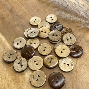 May include: A collection of brown and beige coconut shell buttons with two holes each. The buttons are arranged in a random pattern on a wooden surface.