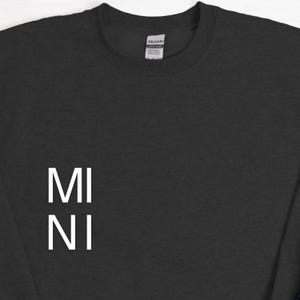 Puede incluir: Una sudadera negra con el texto blanco "MI NI" apilado verticalmente en la parte delantera. La sudadera tiene cuello redondo y mangas largas. La etiqueta de la marca Gildan es visible en la parte posterior del cuello.