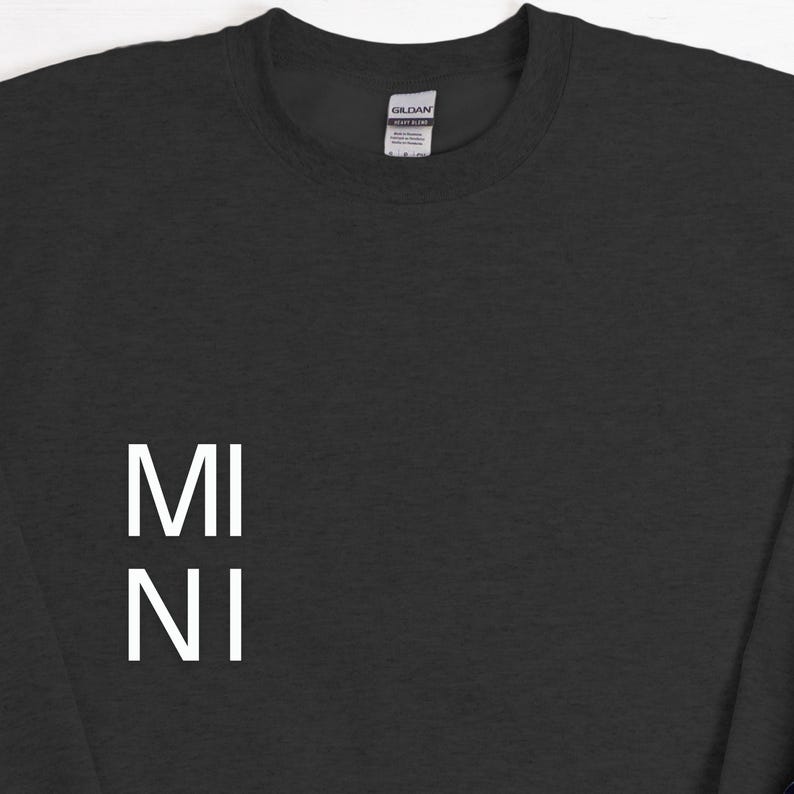 Puede incluir: Una sudadera negra con el texto blanco "MI NI" apilado verticalmente en la parte delantera. La sudadera tiene cuello redondo y mangas largas. La etiqueta de la marca Gildan es visible en la parte posterior del cuello.