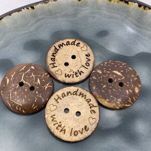 Könnte beinhalten: Drei braune Kokosnuss-Knöpfe mit jeweils zwei Löchern. Die Knöpfe haben den Text "Handmade with love" eingraviert.