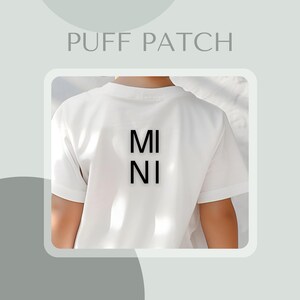 Könnte beinhalten: Weißes T-Shirt mit dem Wort "MINI" in Schwarz auf dem Rücken gedruckt. Das Shirt hat kurze Ärmel und scheint aus einem weichen Material zu sein. Der Text "PUFF PATCH" befindet sich oben im Bild.