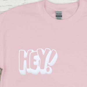 Może przedstawiać: R&oacute;żowy t-shirt z białym nadrukiem "HEY!" z przodu.