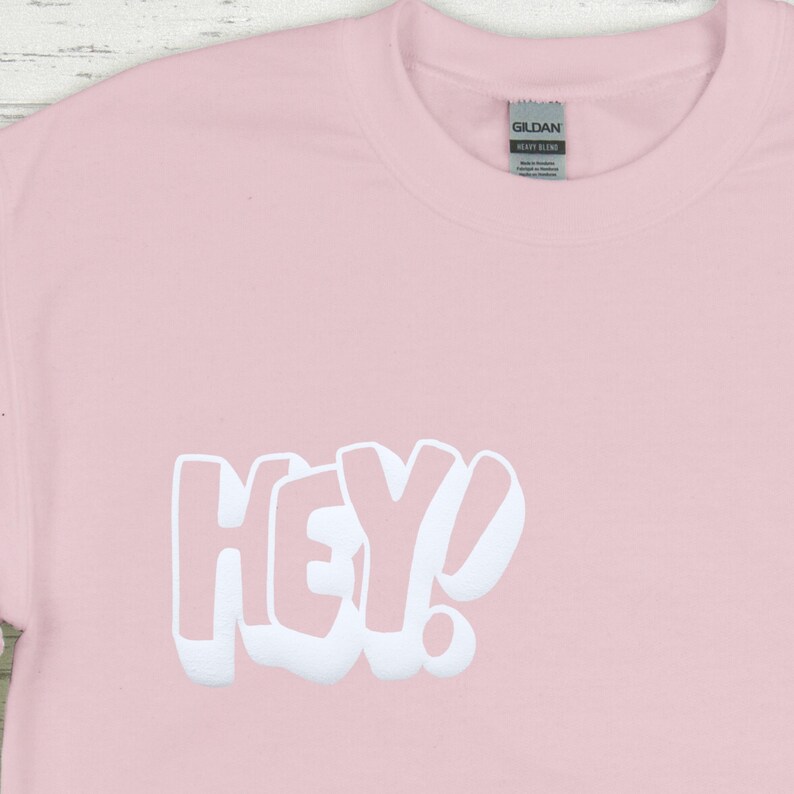 Może przedstawiać: R&oacute;żowy t-shirt z białym nadrukiem "HEY!" z przodu.