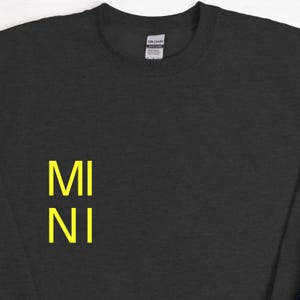 Puede incluir: Una sudadera gris oscuro con la palabra "MINI" impresa verticalmente en letras amarillo brillante en el lado izquierdo. La sudadera tiene cuello redondo y una etiqueta en la parte posterior del cuello.