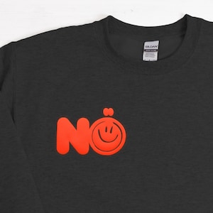 Może przedstawiać: Ciemnoszara bluza z napisem "N&ouml;" w jasnopomarańczowym kolorze, z uśmiechniętą buzią z kropką nad "o". Bluza wykonana jest z miękkiego materiału i jest idealna do noszenia na co dzień.