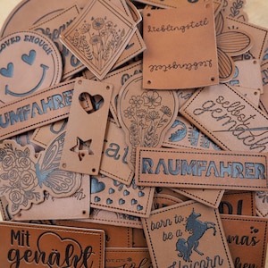 Könnte beinhalten: Eine Sammlung brauner Lederpatches mit verschiedenen Designs und Texten, darunter "Lieblingsteil", "Raumfahrer", "Mit genäht", "Selbst gemacht" und "Born to be a unicorn".