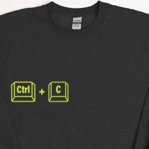 Może przedstawiać: Czarna bluza z neonowo-ż&oacute;łtą grafiką skr&oacute;tu klawiaturowego "Ctrl + C". Tekst i symbole są zamknięte w kwadratowym obrysie. Widoczna jest metka marki Gildan.