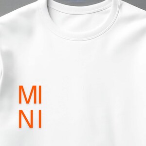 Puede incluir: Una sudadera blanca de cuello redondo con el texto naranja "MI NI" apilado verticalmente en la parte inferior izquierda. La sudadera tiene un dise&ntilde;o simple y limpio.