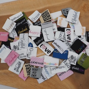 May include: A selection of fabric labels in various colours, including white, pink, black, and blue, with embroidered text. The labels feature words like "happy," "love," and "handmade," arranged on a wooden surface.