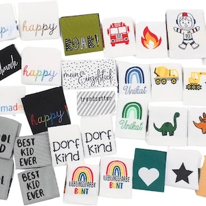 Puede incluir: Una colección de etiquetas de tela con varios diseños y texto. Algunas etiquetas presentan la palabra "happy" en diferentes colores, mientras que otras tienen frases como "Cool Kid" y "Best Kid Ever". También hay etiquetas con ilustraciones de un cohete, un dinosaurio y un camión de bomberos.
