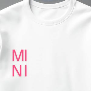 Puede incluir: Sudadera blanca de cuello redondo con la palabra "MINI" impresa verticalmente en letras rosa brillante en la parte delantera. La sudadera tiene un dise&ntilde;o cl&aacute;sico e informal, adecuado para el uso diario.