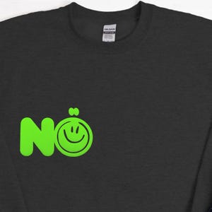 Może przedstawiać: Ciemnoszara bluza z neonowo-zielonym wzorem. Wz&oacute;r przedstawia słowo "N&ouml;" ze znakiem uśmiechu zastępującym "o". Bluza ma okrągły dekolt i długie rękawy.