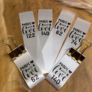 Könnte beinhalten: Fünf weiße Satinband-Etiketten mit schwarzem Text, der "Made with love" und eine Zahl lautet. Die Zahlen auf den Etiketten sind 122, 140, 62, 74 und 62.