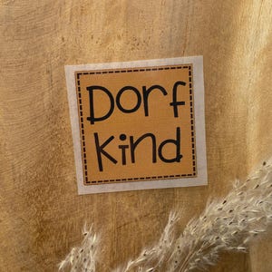 Op de afbeelding: Een vierkante sticker met de woorden "Dorf Kind" in zwart, in een bruine rechthoek met een stippellijn. De sticker bevindt zich op een houten oppervlak, met een takje gedroogd siergras op de voorgrond.