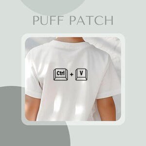 Könnte beinhalten: Weißes T-Shirt mit einer schwarzen Grafik der Ctrl + V-Tasten, darüber der Text "PUFF PATCH". Das T-Shirt hat kurze Ärmel.