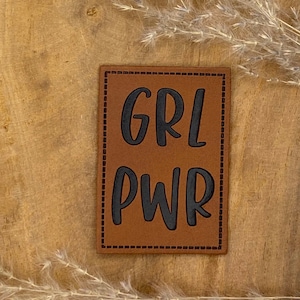 Peut inclure: Patch en cuir marron avec des coutures noires et le texte "GRL PWR" dans une police stylisée.