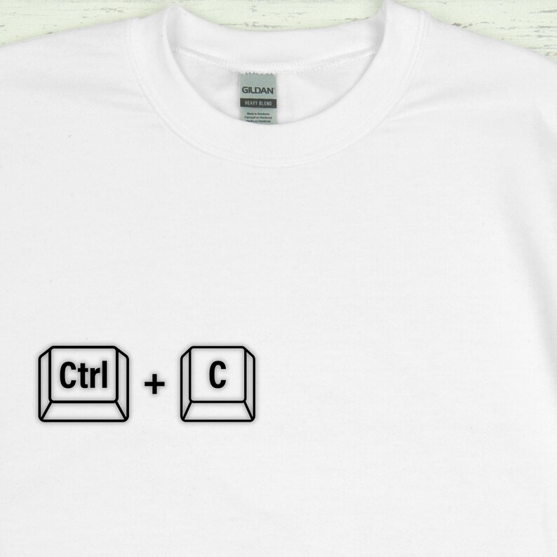 Może przedstawiać: Biała koszulka z czarną grafiką skr&oacute;tu klawiaturowego "Ctrl + C". Koszulka ma okrągły dekolt i jest wykonana z miękkiego, wygodnego materiału. Etykieta Gildan Heavy Blend jest widoczna na dekolcie.