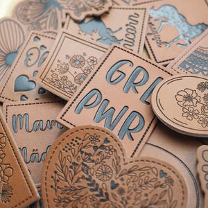 Könnte beinhalten: Eine Sammlung brauner Lederaufnäher mit verschiedenen Designs und Texten. Einige Aufnäher zeigen florale Muster, Herzen und die Wörter "GRL PWR" und "unicorn". Die Aufnäher haben verschiedene Formen, darunter Quadrate, Kreise und Blumen.