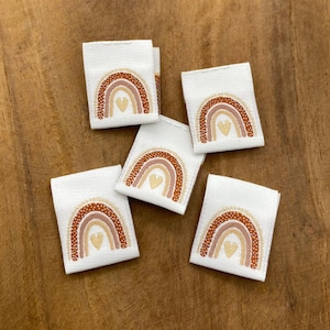 Puede incluir: Cinco etiquetas de tela blanca con un diseño de arcoíris en tonos marrones, naranjas y rosas. Cada arcoíris tiene un pequeño corazón en el centro. Las etiquetas son perfectas para agregar un toque de fantasía a los artículos hechos a mano.