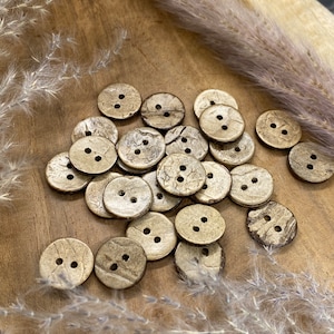 Könnte beinhalten: Eine Sammlung runder, brauner und goldfarbener Knöpfe mit jeweils zwei Löchern. Die Knöpfe sind aus natürlichen Materialien gefertigt und haben eine rustikale, strukturierte Oberfläche.