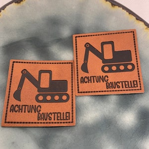 Könnte beinhalten: Zwei braune Leder-Patches mit schwarzen Nähten und einer schwarzen Silhouette eines Baggers. Der Text auf jedem Patch lautet "ACHTUNG BAUSTELLE!"