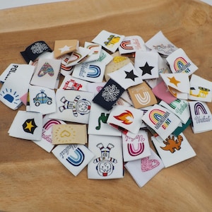 Puede incluir: Una colección de pequeñas etiquetas de tela blanca con varios diseños bordados, incluyendo arcoíris, estrellas y personajes de dibujos animados. Algunas etiquetas presentan texto, como "Unikat" y "Lieblingsfarbe Bunt". Las etiquetas están dispuestas sobre una superficie de madera.