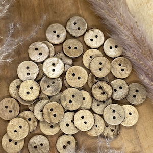 Pode incluir: Um monte de botões de casca de coco natural de cor castanho claro com um brilho dourado. Os botões têm dois furos cada um e estão dispostos em um padrão aleatório sobre uma superfície de madeira.