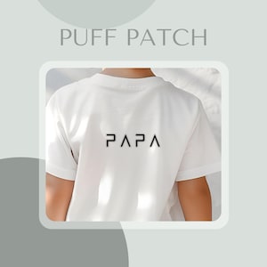 Könnte beinhalten: Weißes T-Shirt mit dem Wort "PAPA" in Schwarz auf dem Rücken gedruckt. Das Shirt hat kurze Ärmel und einen Rundhalsausschnitt. Der Text "PUFF PATCH" steht über dem Shirt.