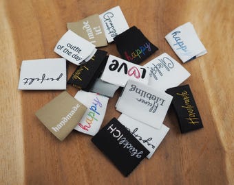 May include: A collection of fabric labels with various phrases and designs, including "handmade", "happy", "outfit of the day", "love", "glücklich", and "perfekt".