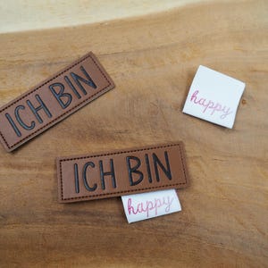以下が含まれることがあります： 黒い文字で「ICH BIN」と書かれた茶色の革のパッチが2つ。ピンクの文字で「happy」と書かれた白いパッチが2つ。