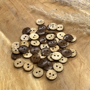 May include: A pile of brown and natural coconut shell buttons with two holes each. The buttons are on a wooden surface.