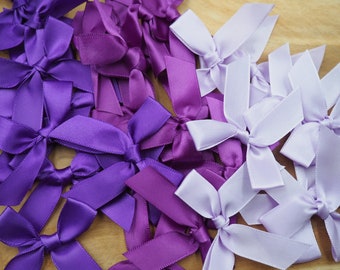 Può includere: Una collezione di fiocchi di nastro di raso viola e lavanda. I fiocchi sono disposti in una pila su una superficie di legno.