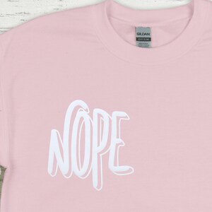 Op de afbeelding: Een roze T-shirt met het woord "NOPE" in wit op de voorkant.