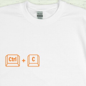 Może przedstawiać: Biała koszulka z okrągłym dekoltem z pomarańczową grafiką klawiszy Ctrl + C. Koszulka wykonana jest z miękkiego materiału i posiada metkę Gildan przy dekolcie. Prosty design dla użytkownik&oacute;w komputer&oacute;w.