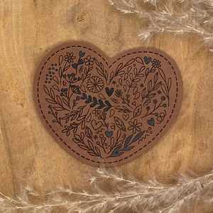 Puede incluir: Un posavasos de cuero marrón en forma de corazón con un diseño floral negro. El posavasos presenta una variedad de flores, hojas y corazones.