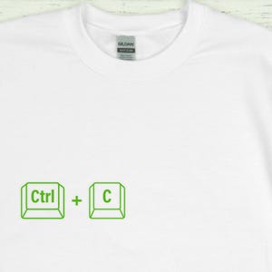 Może przedstawiać: Biała koszulka z okrągłym dekoltem z zieloną grafiką skr&oacute;tu klawiaturowego Ctrl + C. Tekst i symbole są w prostym, obrysowanym stylu. Koszulka ma metkę z tyłu szyi.