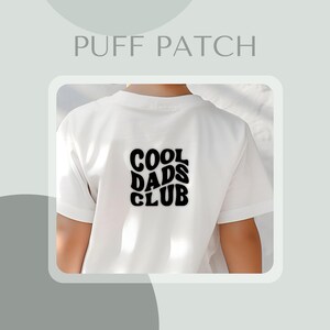 May include: White t-shirt featuring a black "COOL DADS CLUB" puff patch on the back. The text is in a retro, wavy font. The shirt has short sleeves.