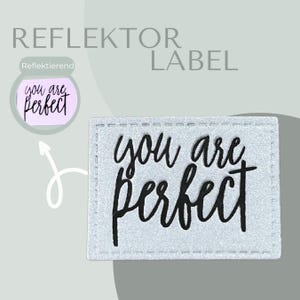 Könnte beinhalten: Ein silbernes reflektierendes Etikett mit dem Text "you are perfect" in schwarzer Kursivschrift. Das Etikett hat einen genähten Rand.