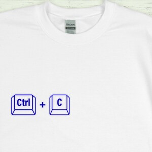 Może przedstawiać: Biała koszulka z niebieską grafiką skr&oacute;tu klawiaturowego "Ctrl + C". Koszulka ma okrągły dekolt i metkę z napisem "Gildan Heavy Blend".