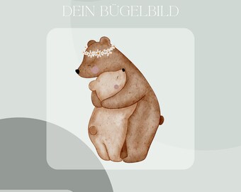 Op de afbeelding: Een strijkapplicatie met een aquarelillustratie van een grote bruine beer met een witte madeliefjeskrans die een kleinere lichtbeige beer omhelst. De tekst DEIN BÜGELBILD staat erboven en Inklusive Anleitung und Test-Bügelbild staat eronder.