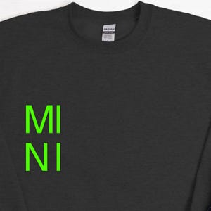 Puede incluir: Sudadera gris oscuro con la palabra "MI NI" en letras de bloque verde brillante. La sudadera tiene cuello redondo y una etiqueta en la parte posterior del cuello.