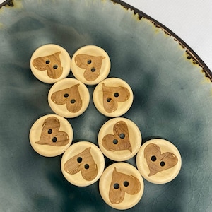 Könnte beinhalten: Acht Holz-Knöpfe mit Herz-Design. Die Knöpfe sind hellbraun mit einem dunkelbraunen Herz-Design. Die Knöpfe sind in einem Kreis auf einem blauen und braunen Teller angeordnet.