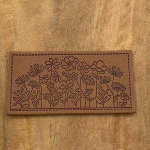 Puede incluir: Un parche de cuero marrón con un borde cosido y un diseño de un campo de flores en negro.