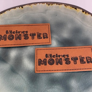 Könnte beinhalten: Zwei braune Leder-Etiketten mit genähten Kanten. Jedes Etikett trägt den Text "Kleines Monster" in schwarzer Schrift mit einem stilisierten Monster-Design.