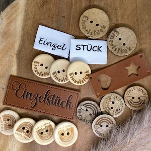 Puede incluir: Una colección de botones de madera con el texto "Einzelstick" en ellos. Los botones son de varias formas y tamaños, incluyendo círculos, estrellas e arcoíris. Algunos de los botones tienen un diseño de corazón. Los botones están sobre una superficie de madera.