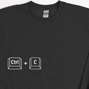 Może przedstawiać: Czarna bluza z białym nadrukiem skr&oacute;tu klawiszowego "Ctrl + C". Bluza marki Gildan ma minimalistyczny design. Idealna dla entuzjast&oacute;w technologii lub fan&oacute;w skr&oacute;t&oacute;w klawiszowych.