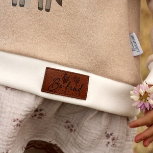 May include: A beige sweatshirt with a white cuff and a brown leather patch that says "Be Kind". The sweatshirt has a white and brown floral print on the bottom.