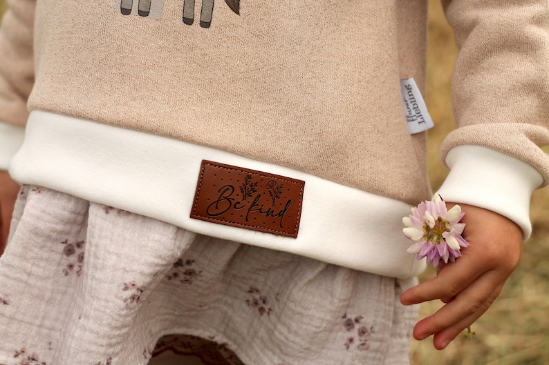 May include: A beige sweatshirt with a white cuff and a brown leather patch that says "Be Kind". The sweatshirt has a white and brown floral print on the bottom.